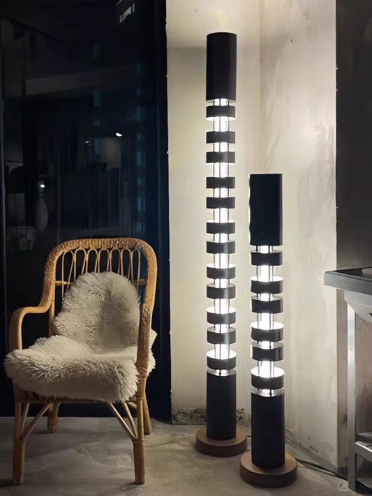 Two modern black floor lamps with illuminated cylindrical columns and circular wooden bases, shown in a styled interior next to a wicker chair with a white faux-fur cushion.