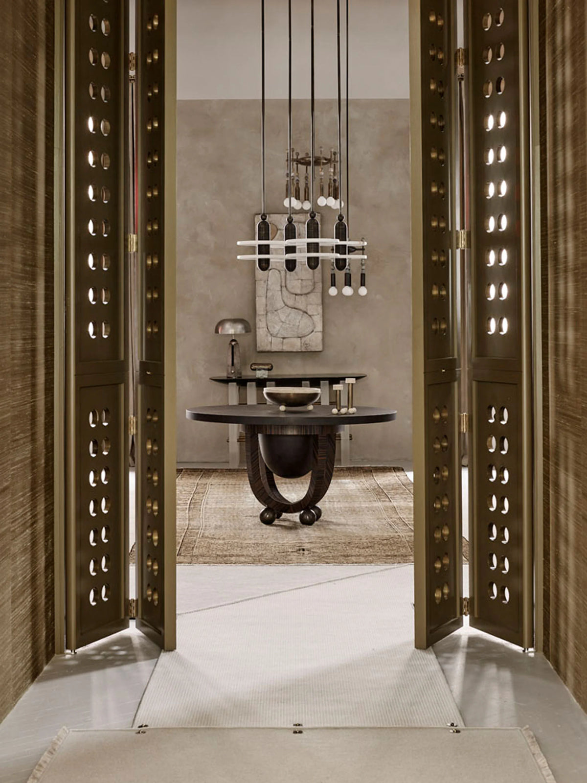 An interior architectural scene viewed through open ornate bronze-toned doors, showing a dining area with a round dark table, pendant lighting, textured wall, and rug.