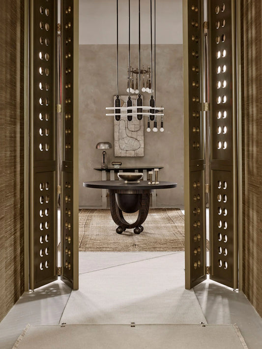 An interior architectural scene viewed through open ornate bronze-toned doors, showing a dining area with a round dark table, pendant lighting, textured wall, and rug.