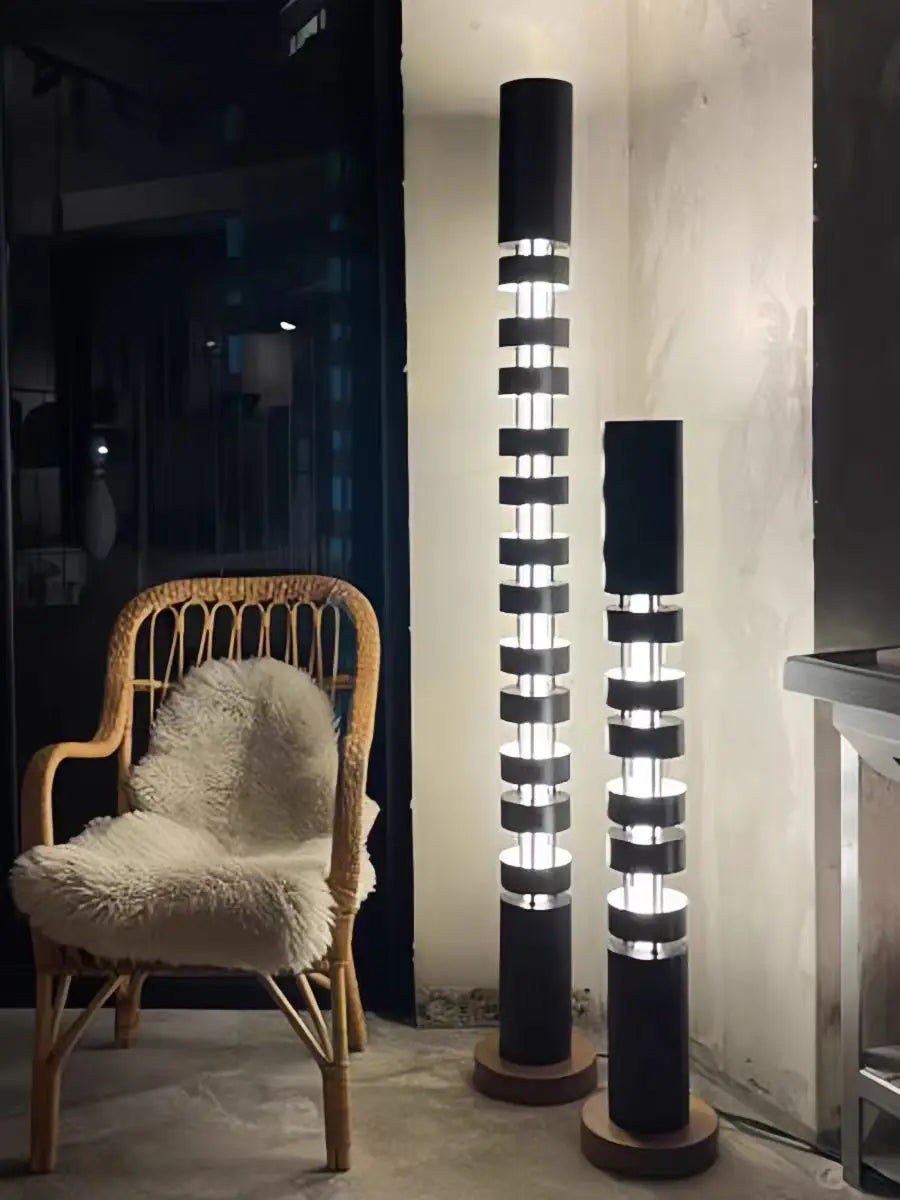 Two modern black floor lamps with illuminated cylindrical columns and circular wooden bases, shown in a styled interior next to a wicker chair with a white faux-fur cushion.