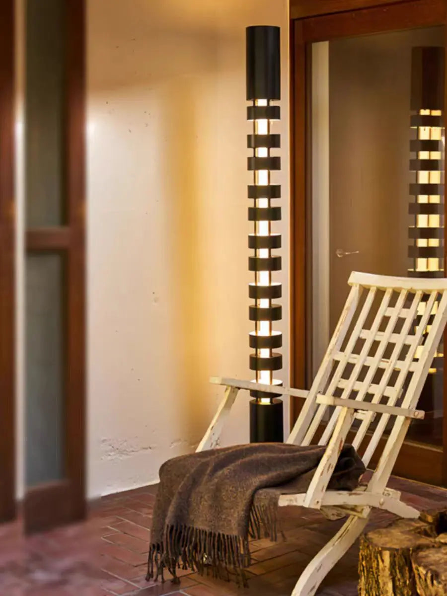 A modern outdoor floor lamp with a black cylindrical base and horizontal slatted shade, glowing with warm light next to a wooden folding chair with a throw blanket.